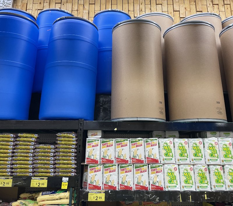 Blue plastic and cardboard shipping barrels displayed above Caribbean grocery staples